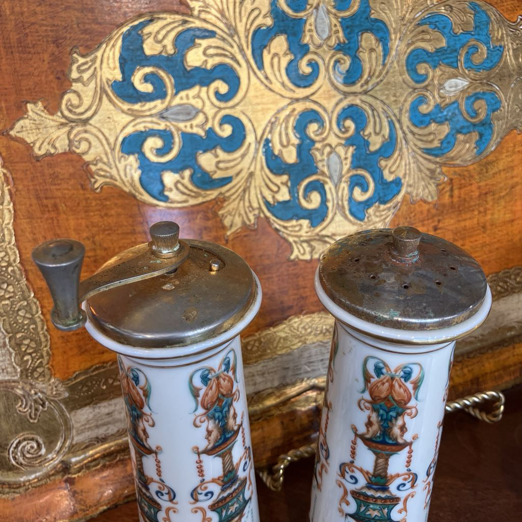Two vintage porcelain pepper mills with silver metal lids and handles, decorated with blue and gold scrollwork, set against an ornate wooden background.