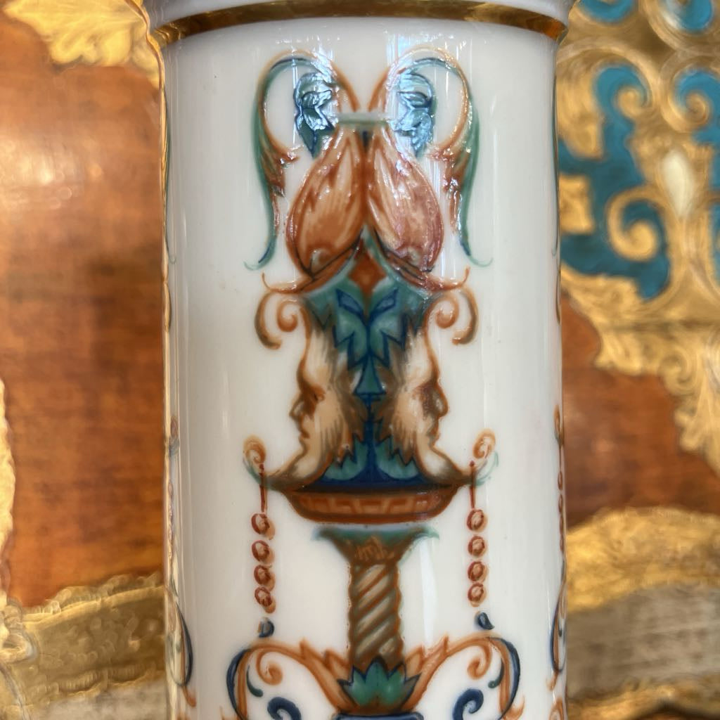 Close-up of a white porcelain vase featuring a hand-painted classical urn motif with gold, teal, and rust-brown details, set against a blurred ornate background.