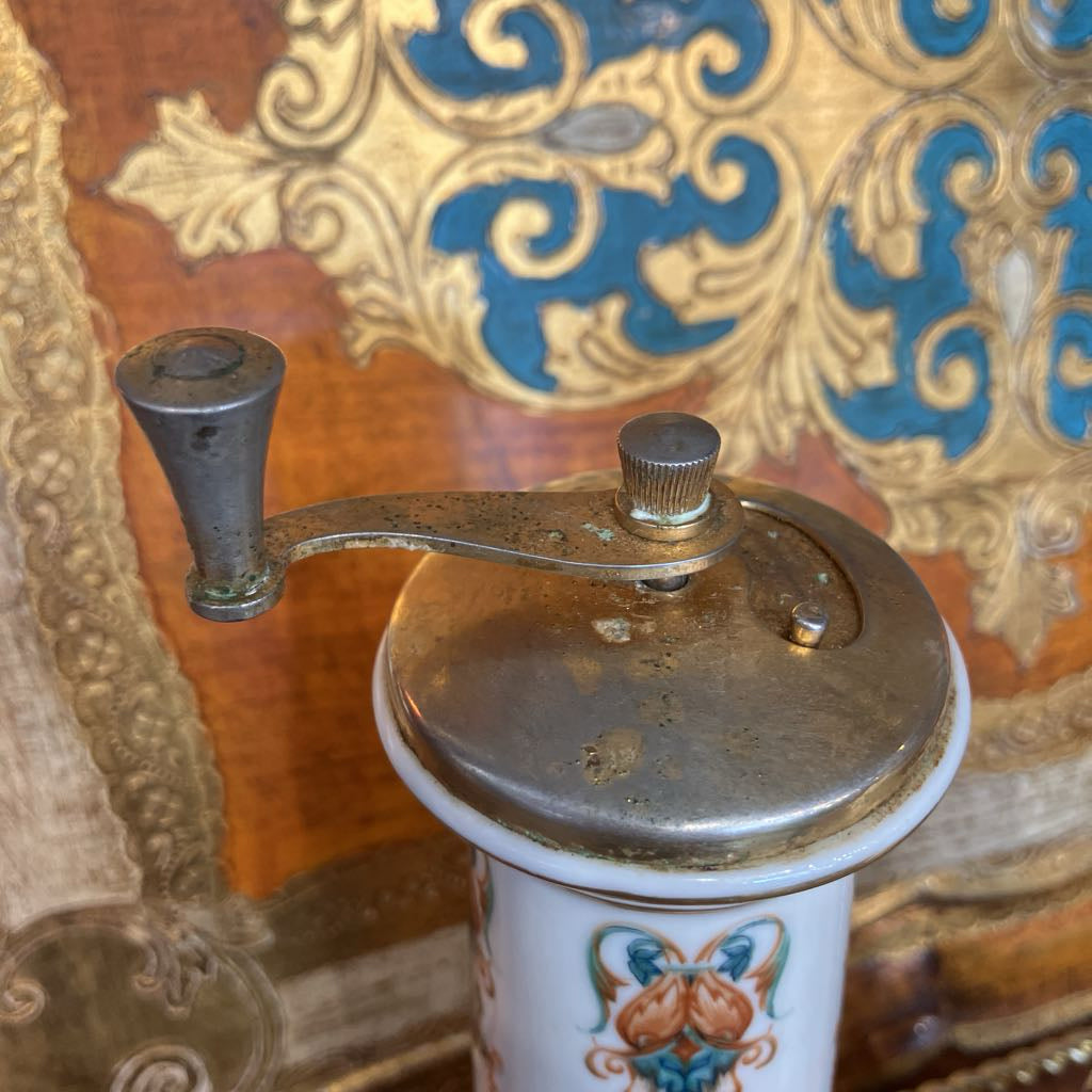 A vintage manual coffee grinder with a metal handle and lid, mounted on a white ceramic body decorated with a colorful butterfly motif, set against an ornate gold and blue patterned background.