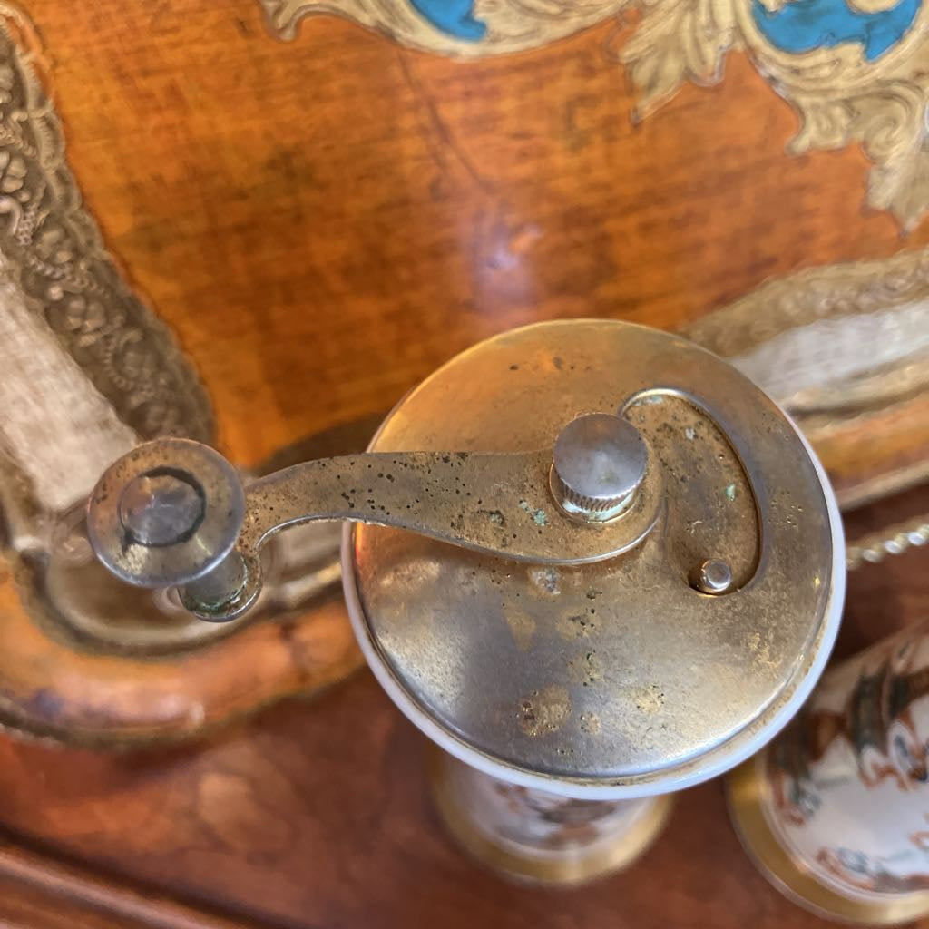 Close-up of a vintage brass or bronze hand-crank grinder resting on an ornate wooden surface with painted details.