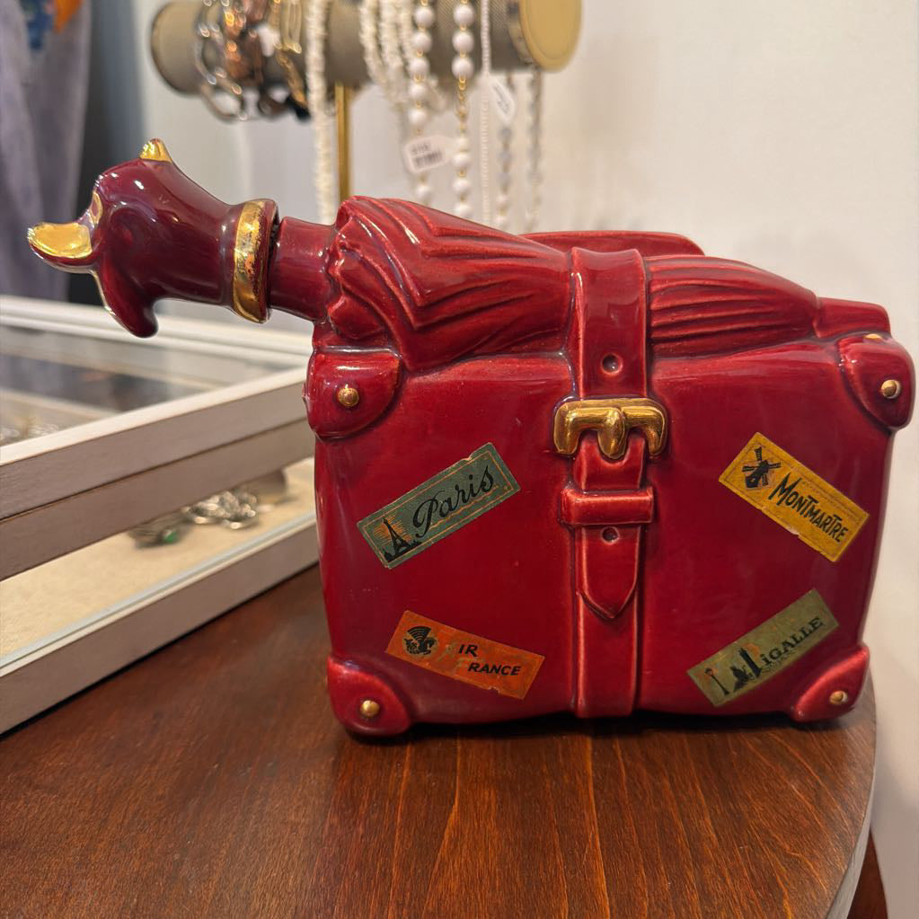 A glossy red ceramic decorative trunk shaped like a suitcase with a horn-like protruding handle on the left, gold-tone hardware, and several faux travel stickers, displayed on a wooden surface.