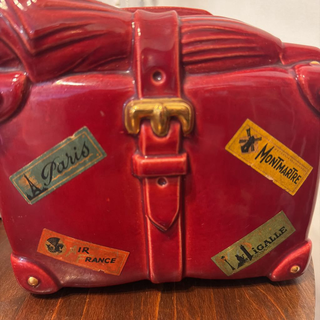 A red ceramic souvenir luggage figurine shaped like a vintage suitcase with a gold-tone buckle and several colorful travel stickers reading Paris, Montmartre, Nice, and Lille.