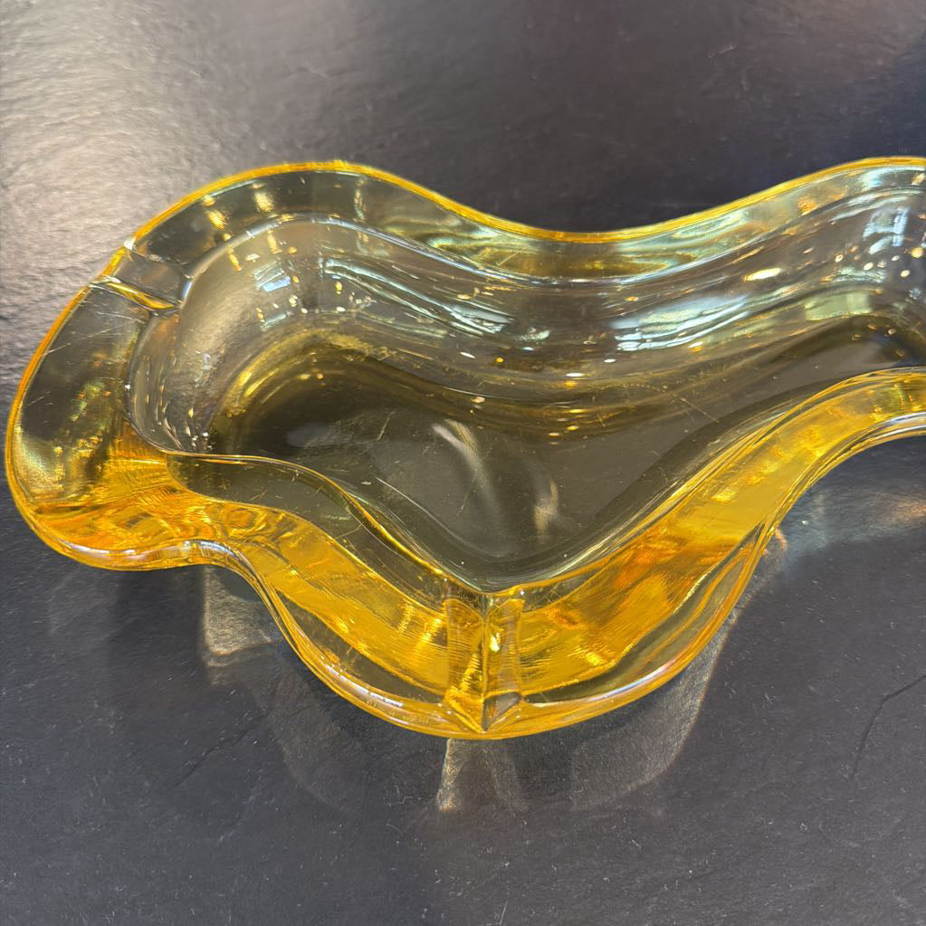 A translucent amber yellow glass ashtray with a wavy, organic shape, photographed on a dark gray surface.