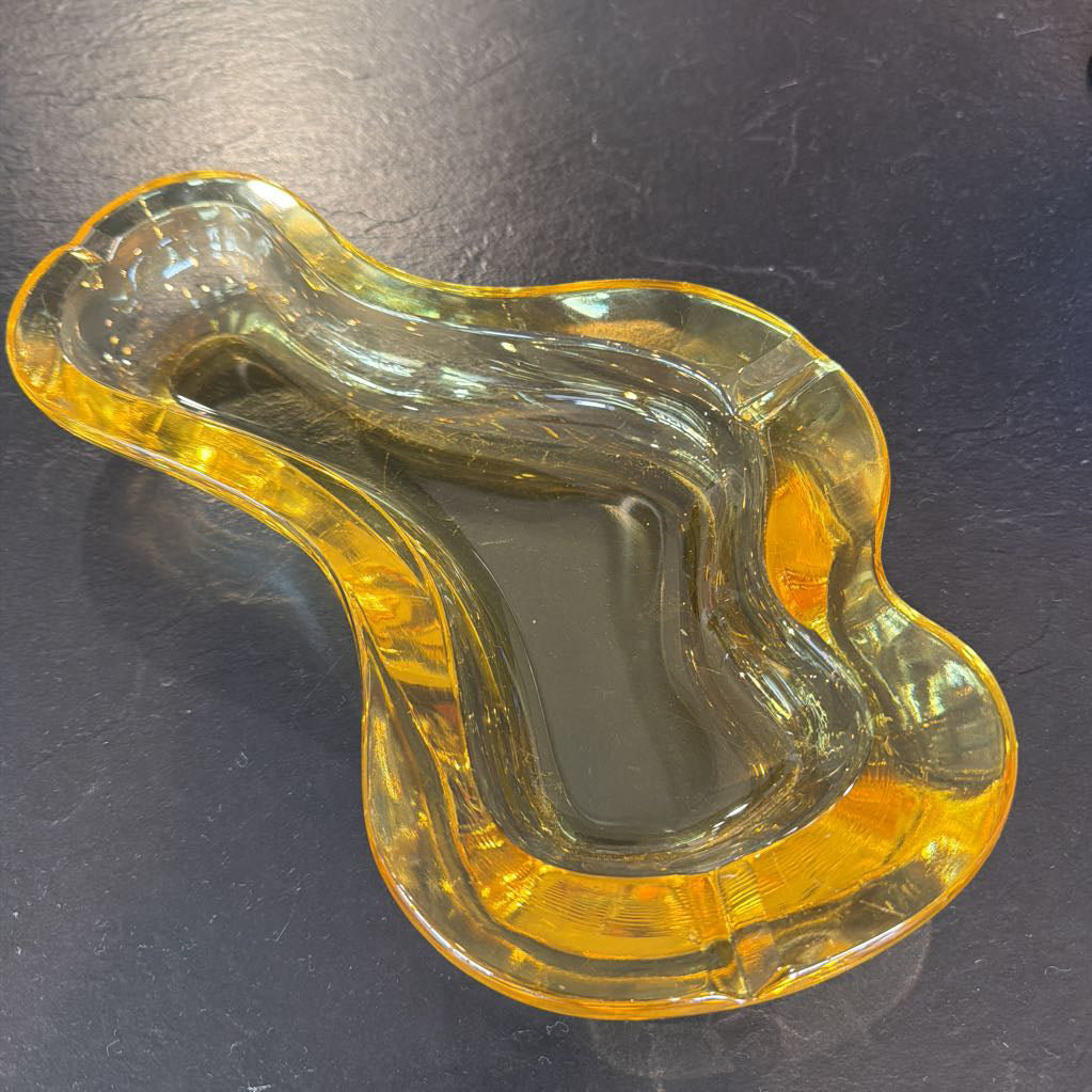 A translucent amber-yellow glass dish with an organic, wavy shape, photographed on a dark textured surface.