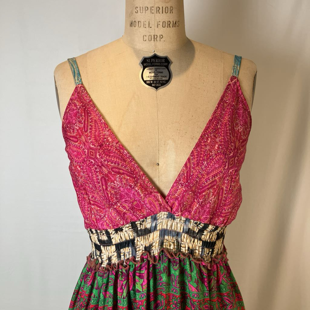 A sleeveless V-neck dress displayed on a vintage wooden mannequin torso. The dress features a bright pink patterned bodice with thin straps, a black, beige, and white patterned waistband, and a green, pink, and black patterned skirt.