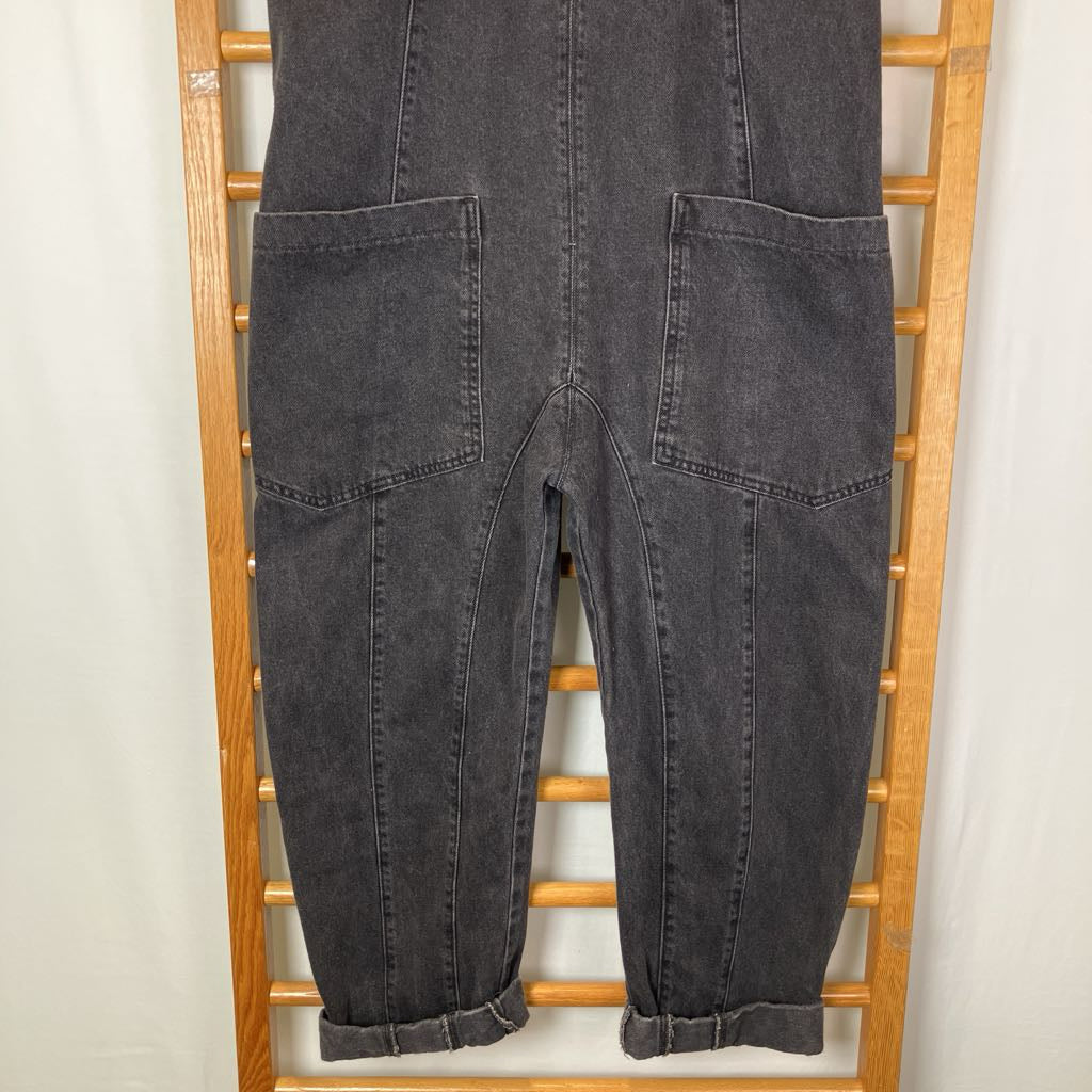 Dark gray washed denim overalls with large front patch pockets, shown hanging on a wooden ladder against a light off-white background.
