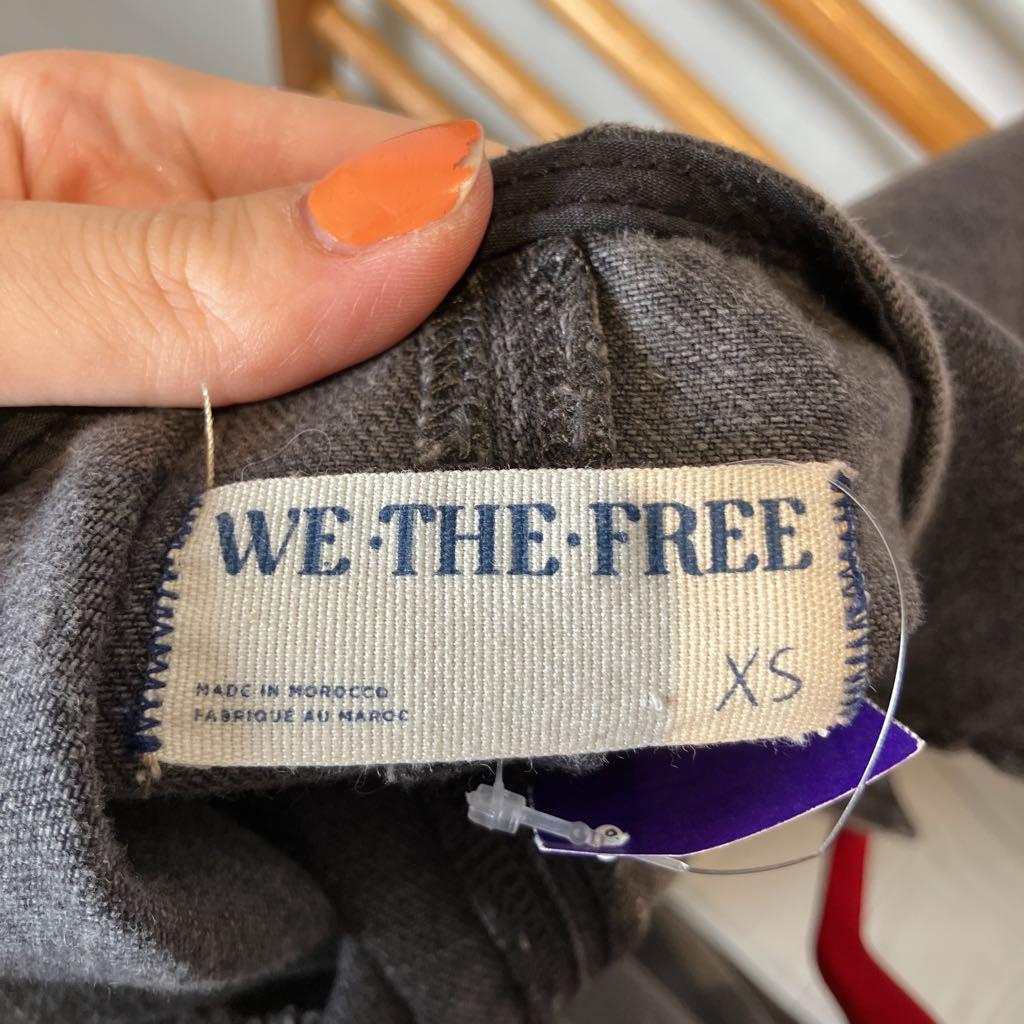A close-up of a hand holding the collar of a dark gray knit garment, showing the sewn-in label that reads WE THE FREE, size XS, with a purple hangtag attached.