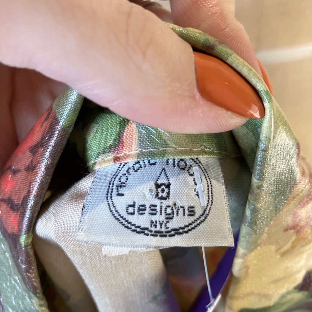 A close-up of a hand holding the collar of a floral satin blouse, showing the brand label 'Nordic House designs NYC'. The blouse features a multicolor floral pattern with green, gold, and red tones.