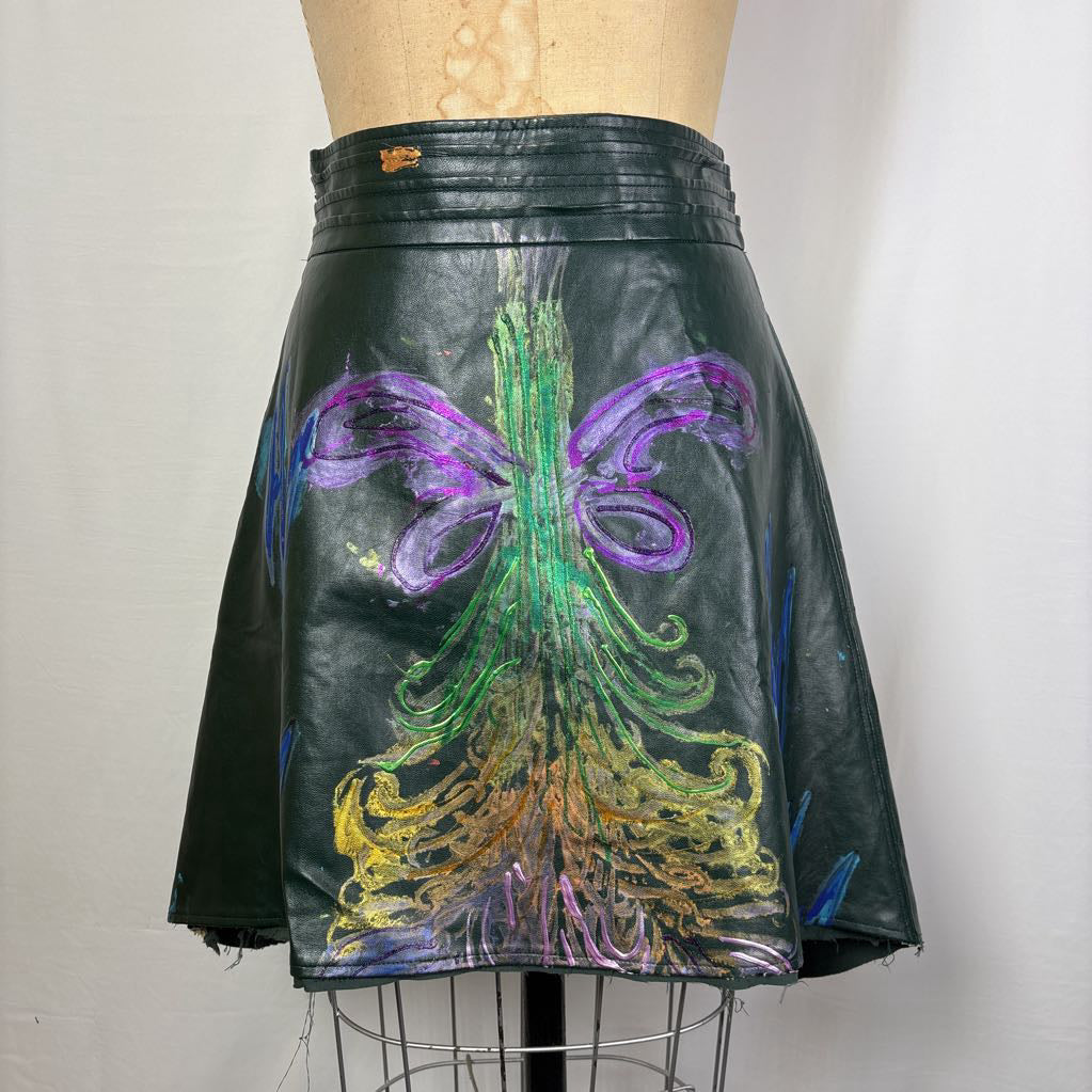 A close-up of a black leather garment, likely a skirt or jacket, featuring a large, vibrant hand-painted design on the back. The artwork depicts a stylized tree with green trunk and branches, purple wings, and gold roots, set against a plain light background.
