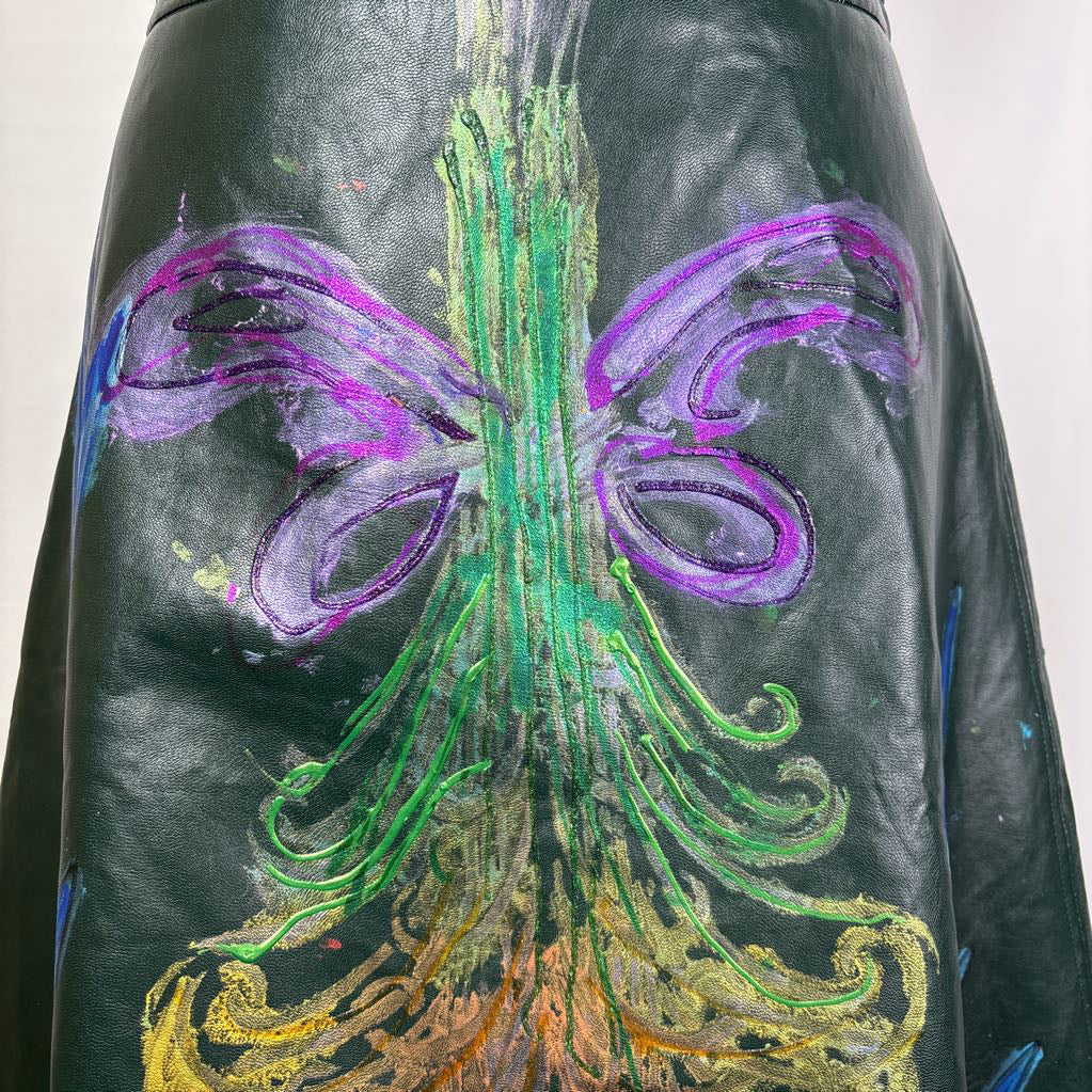 A close-up of a black leather garment, likely a skirt or jacket, featuring a large, vibrant hand-painted design on the back. The artwork depicts a stylized tree with green trunk and branches, purple wings, and gold roots, set against a plain light background.