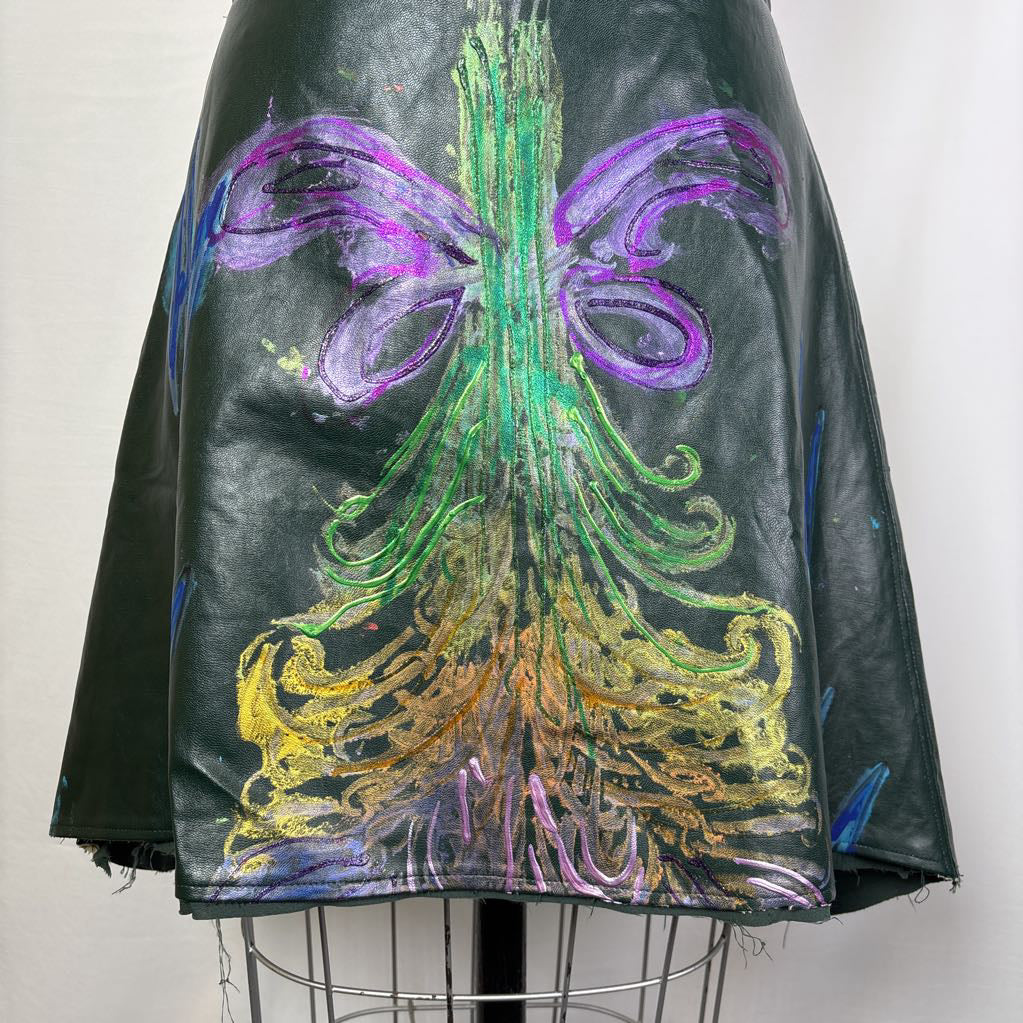 A close-up of a black leather garment, likely a skirt or jacket, featuring a large, vibrant hand-painted design on the back. The artwork depicts a stylized tree with green trunk and branches, purple wings, and gold roots, set against a plain light background.