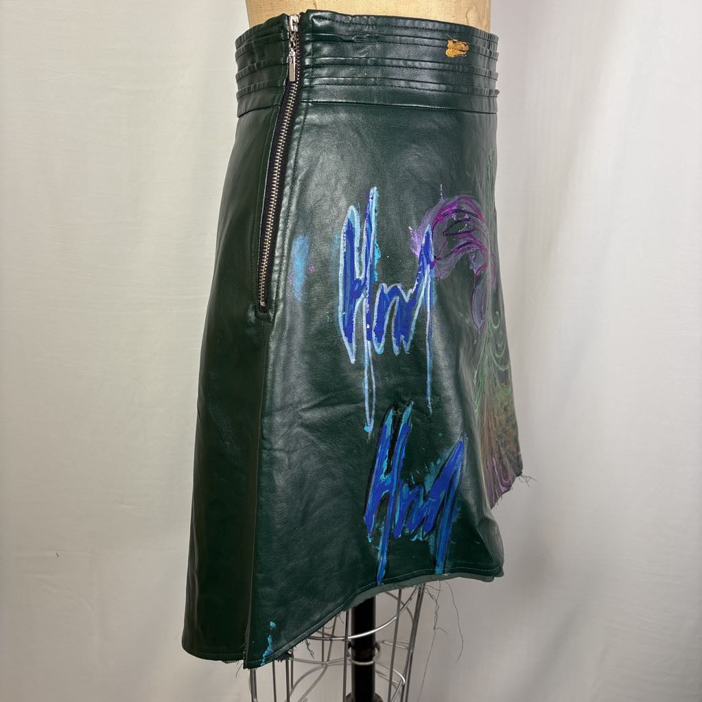 Side close-up of a black leather garment, likely a skirt or jacket, featuring a large, vibrant hand-painted design on the back. The artwork depicts a stylized tree with green trunk and branches, purple wings, and gold roots, set against a plain light background.