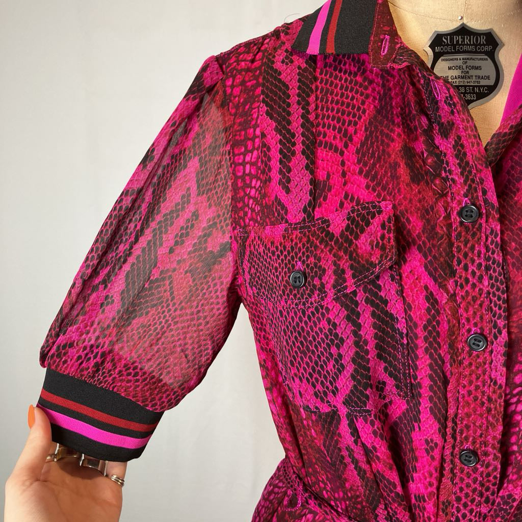 A close-up of a pink and black snakeskin-patterned blouse on a mannequin, with a hand holding the sleeve cuff to show the striped trim. The collar label reads 'SUPERBLO' and 'MILANO, ITALY'.