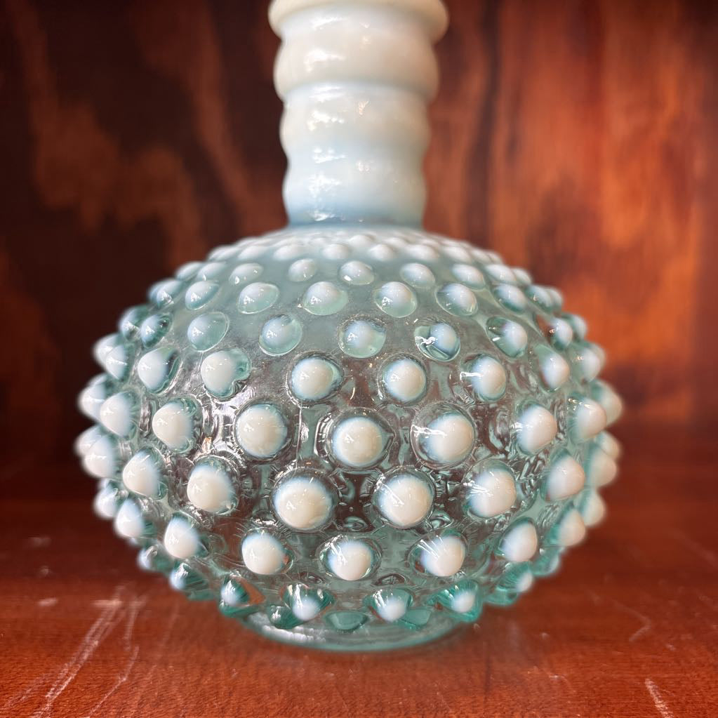 A clear and milk hobnail glass perfume bottle with a stopper in a wooden cabinet.