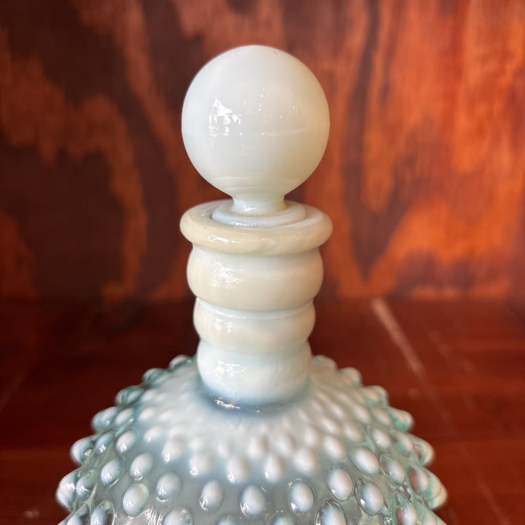 A clear and milk hobnail glass perfume bottle with a stopper in a wooden cabinet.