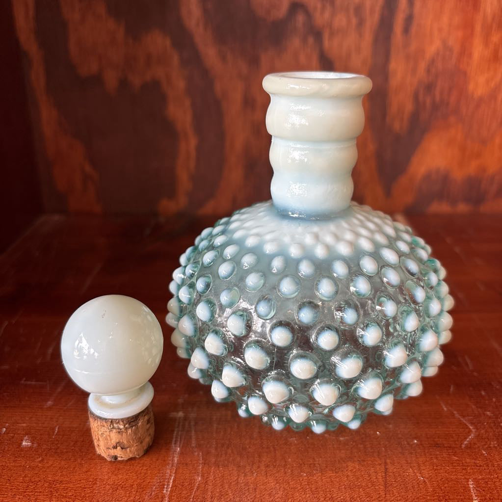A clear and milk hobnail glass perfume bottle with a stopper in a wooden cabinet.