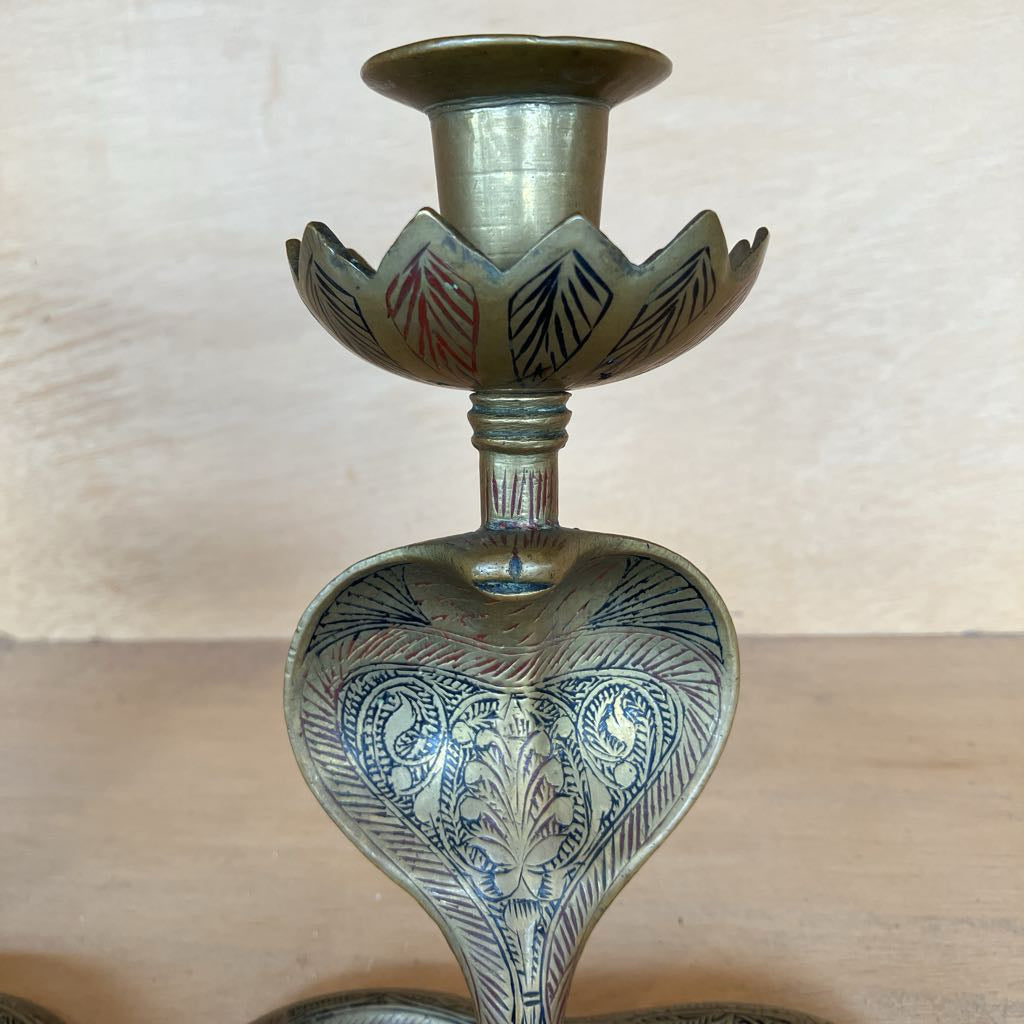 An ornate brass candlestick holder with hand-painted blue, red, and black floral patterns on a light wood surface against a beige background.