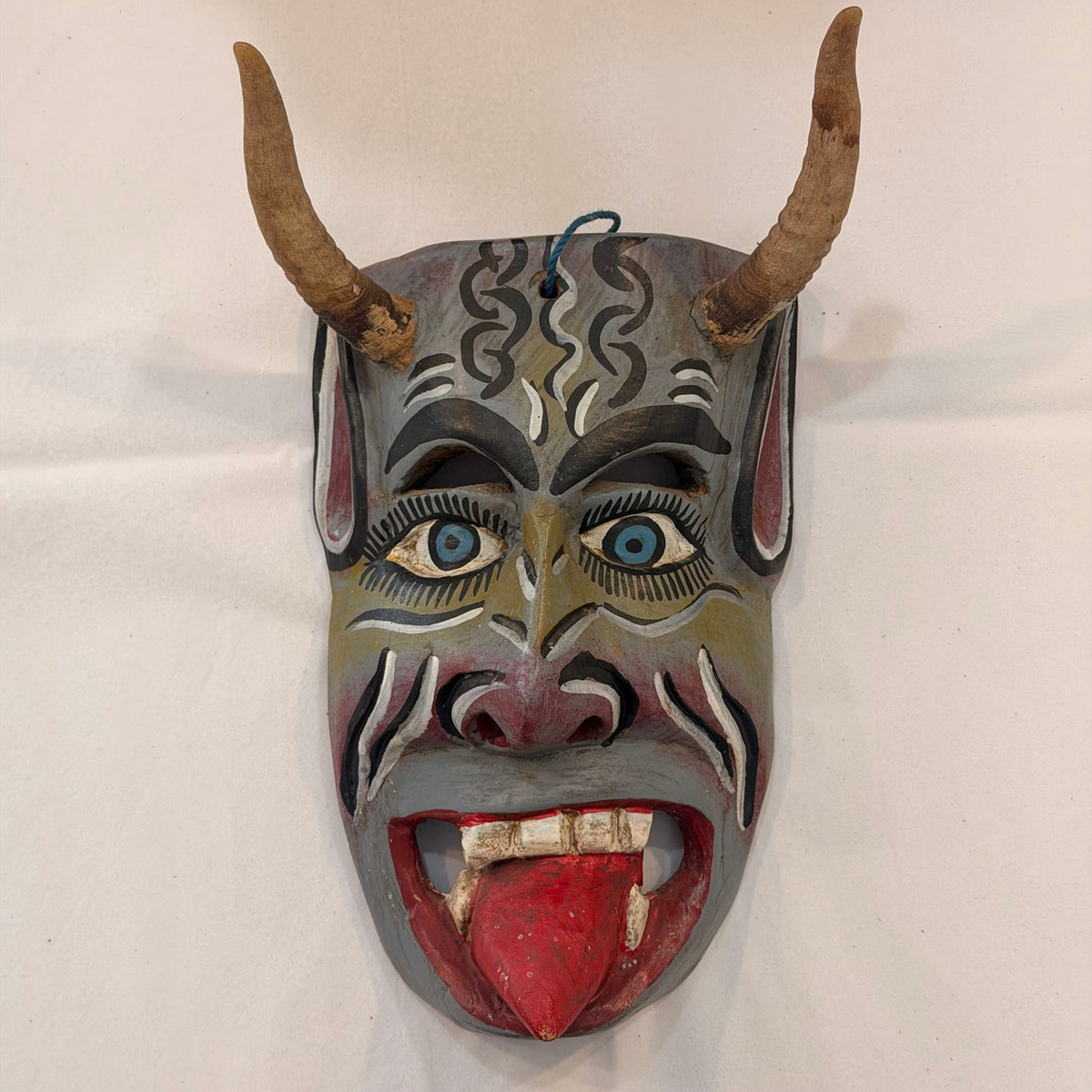 Hand-carved wooden mask with horns, featuring a stylized face with blue eyes, white and black patterns, and a red tongue, mounted against a plain white wall.