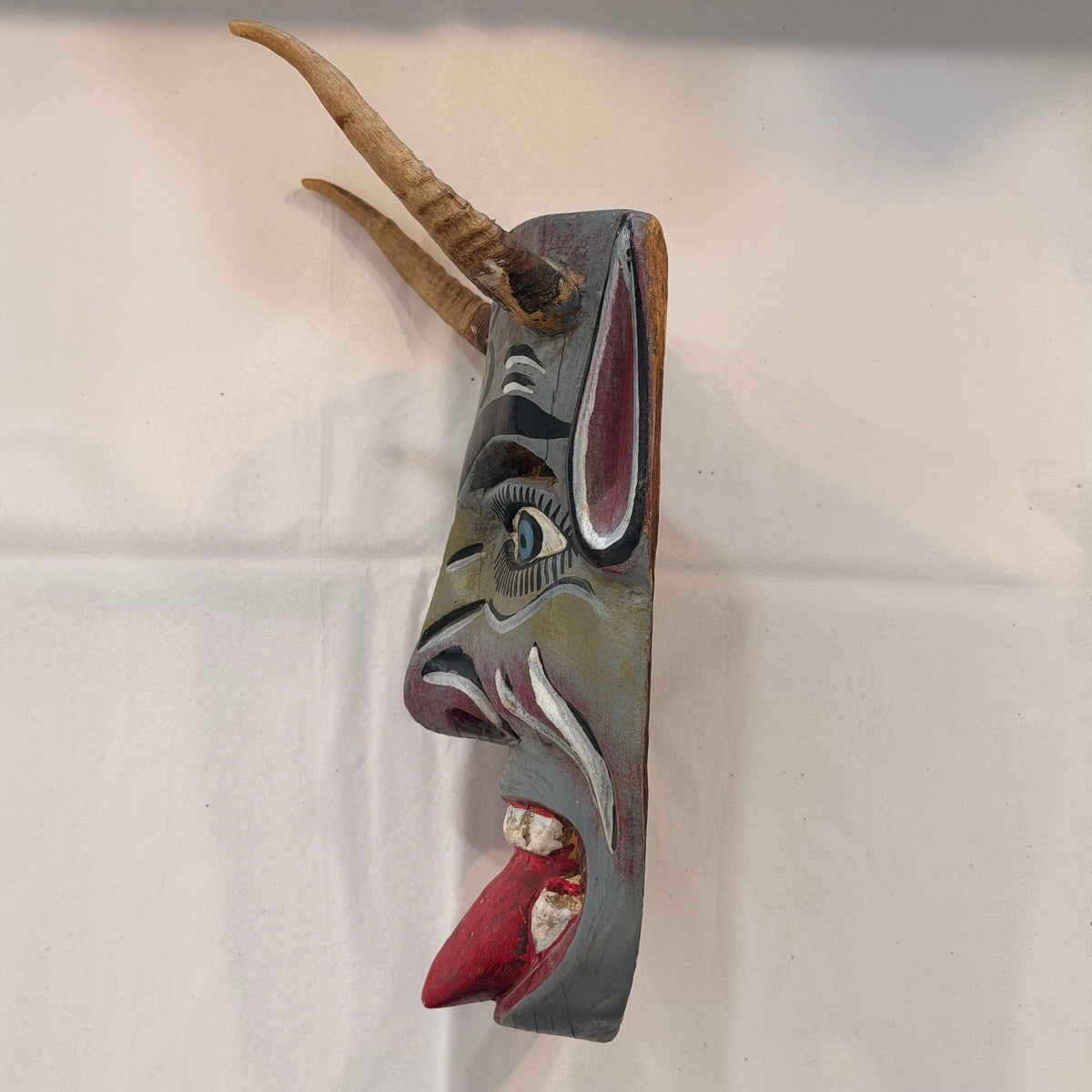A hand-carved wooden mask with antler-like horns, featuring a stylized face with red, white, black, and gray painted details, displayed against a light fabric background.
