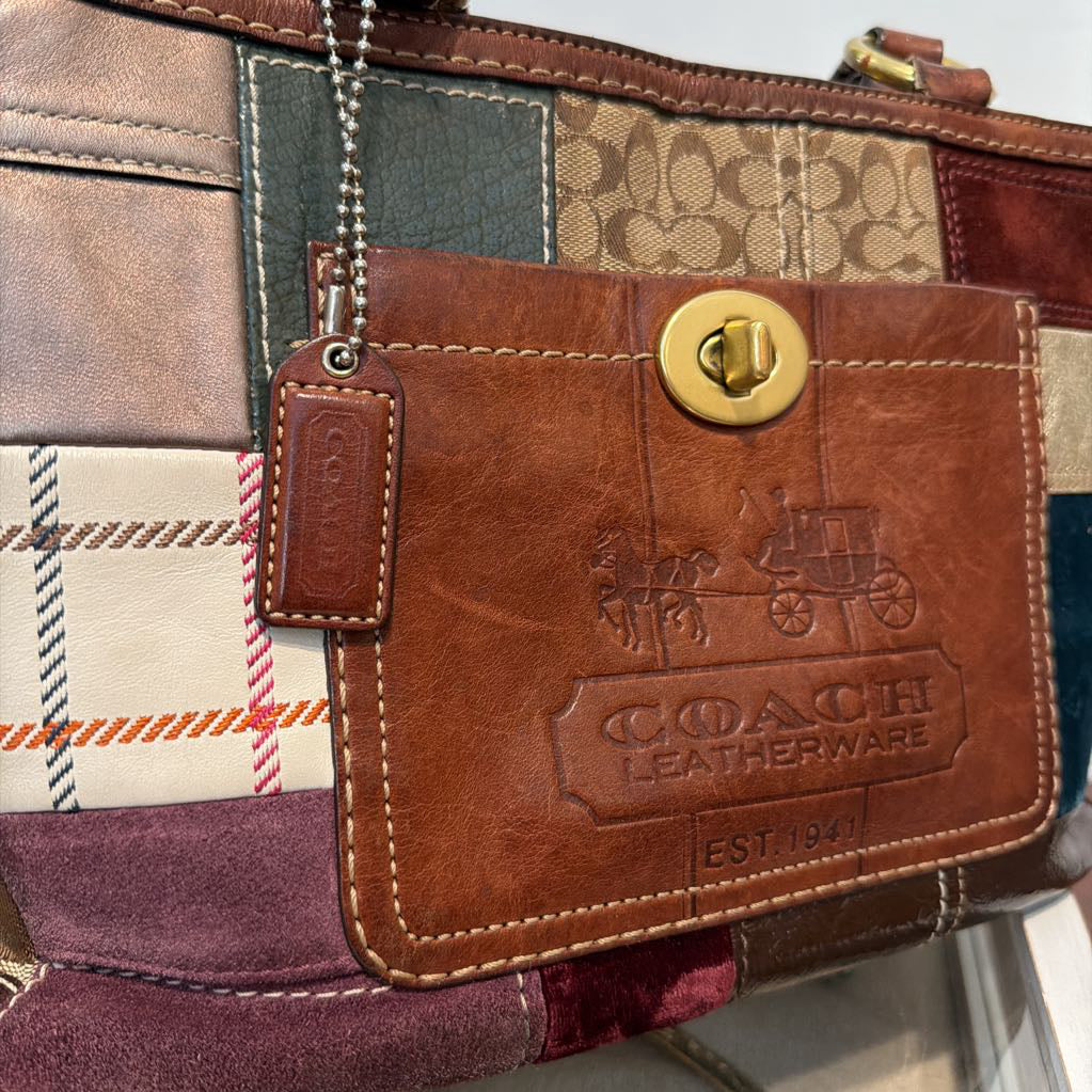 Close-up of a Coach leather wallet or card holder with a brown embossed leather patch, gold-tone turn-lock closure, and a hanging leather tag on a patchwork bag background.