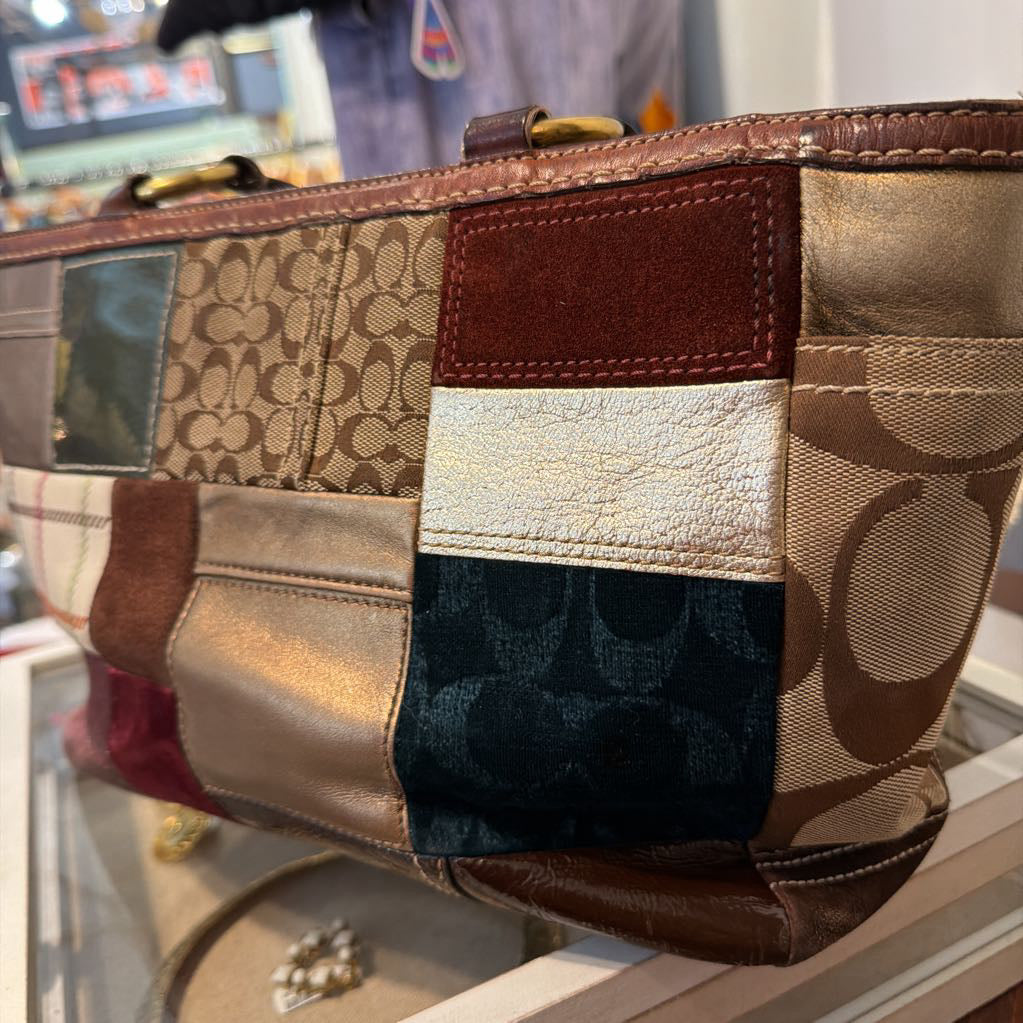 A close-up of a Coach patchwork handbag featuring multiple panels of signature monogram canvas in beige and brown, metallic gold leather, dark navy monogram fabric, and a reddish-brown suede section.