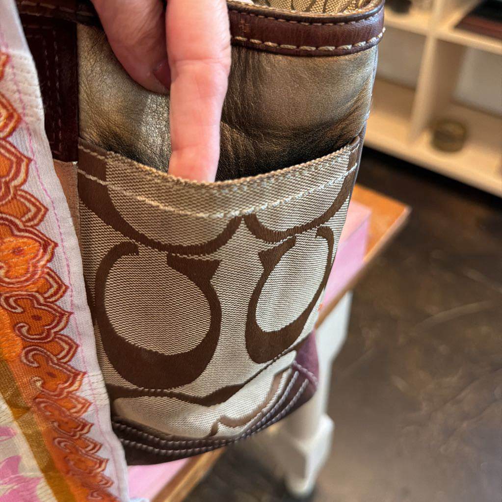 Close-up of a hand holding a Coach tote bag, showing the signature monogram canvas pattern and brown leather trim in a shop setting.