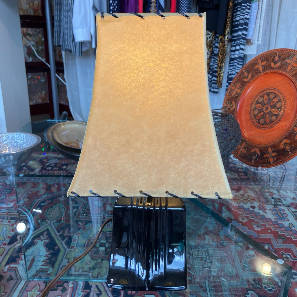 A table lamp with a large, warm beige fabric shade and a glossy black base, displayed on a glass table in a busy shop setting.