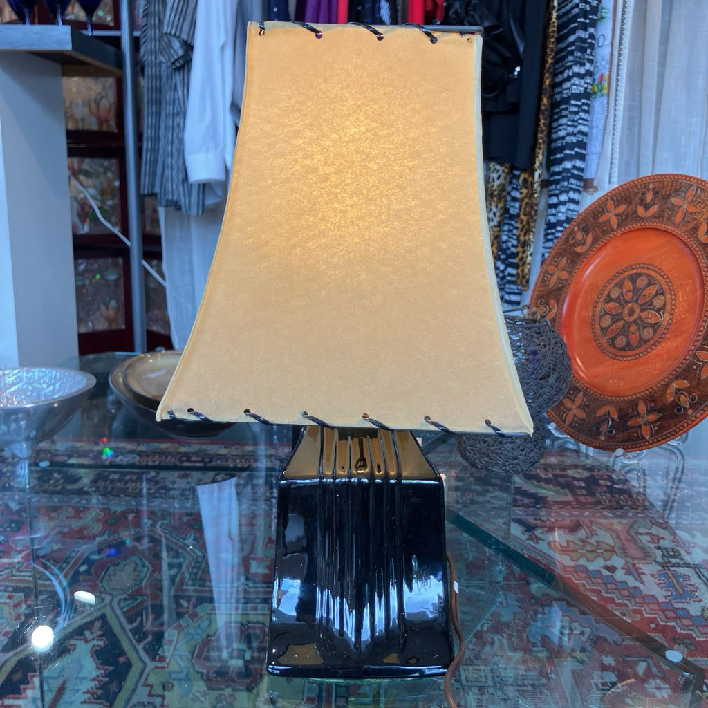 A table lamp with a textured beige fabric shade and a glossy black fluted base, displayed on a glass table in a shop setting.