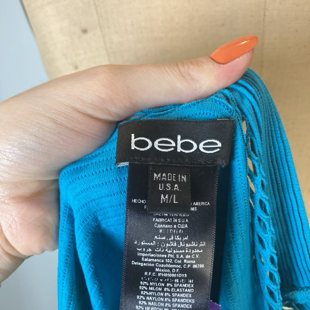 A close-up of a hand holding a bright turquoise blue knit garment, showing the black fabric brand label that reads 'bebe'.