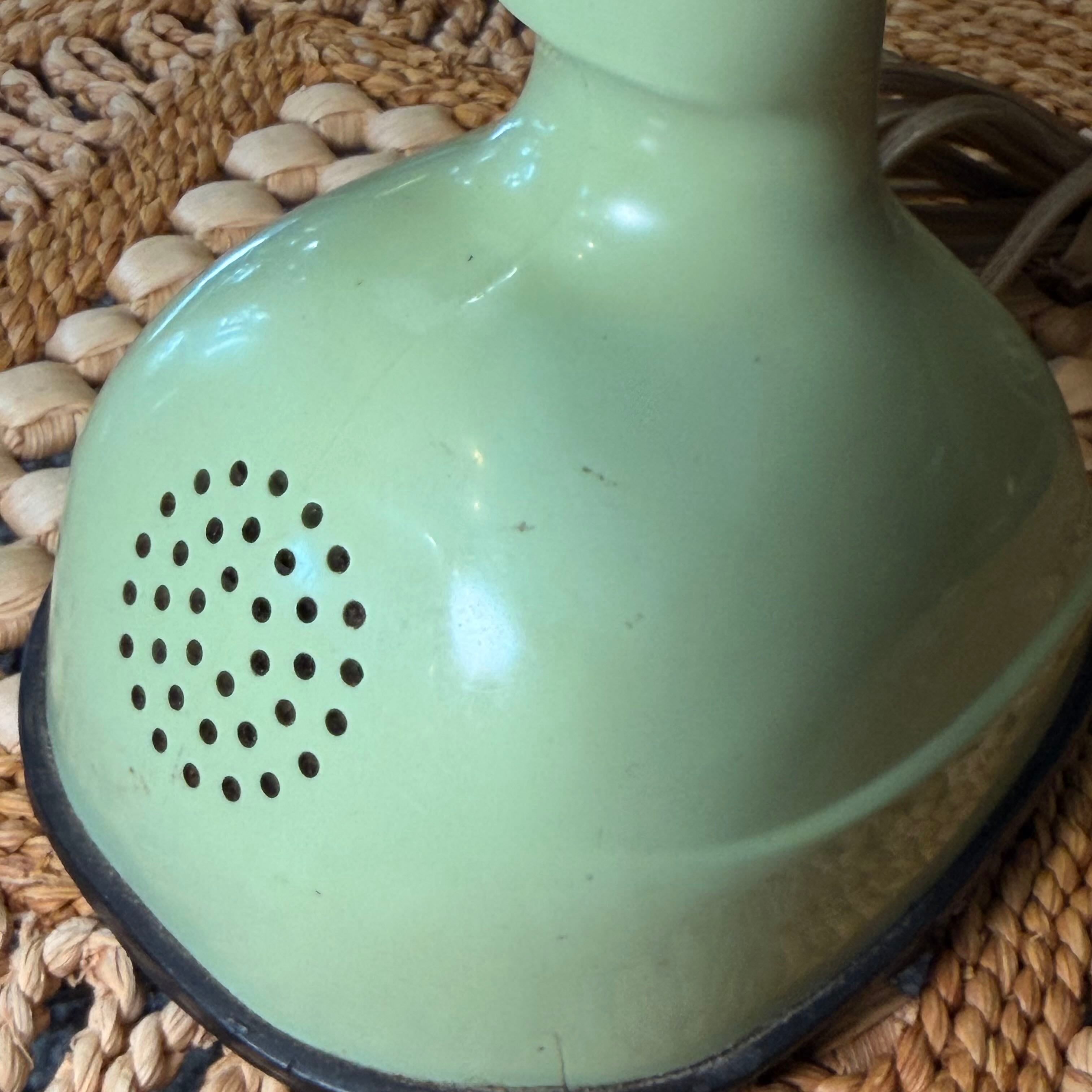 A close-up of a vintage light green enamel hair dryer base with a circular vent and a black cord, resting on a woven straw mat.