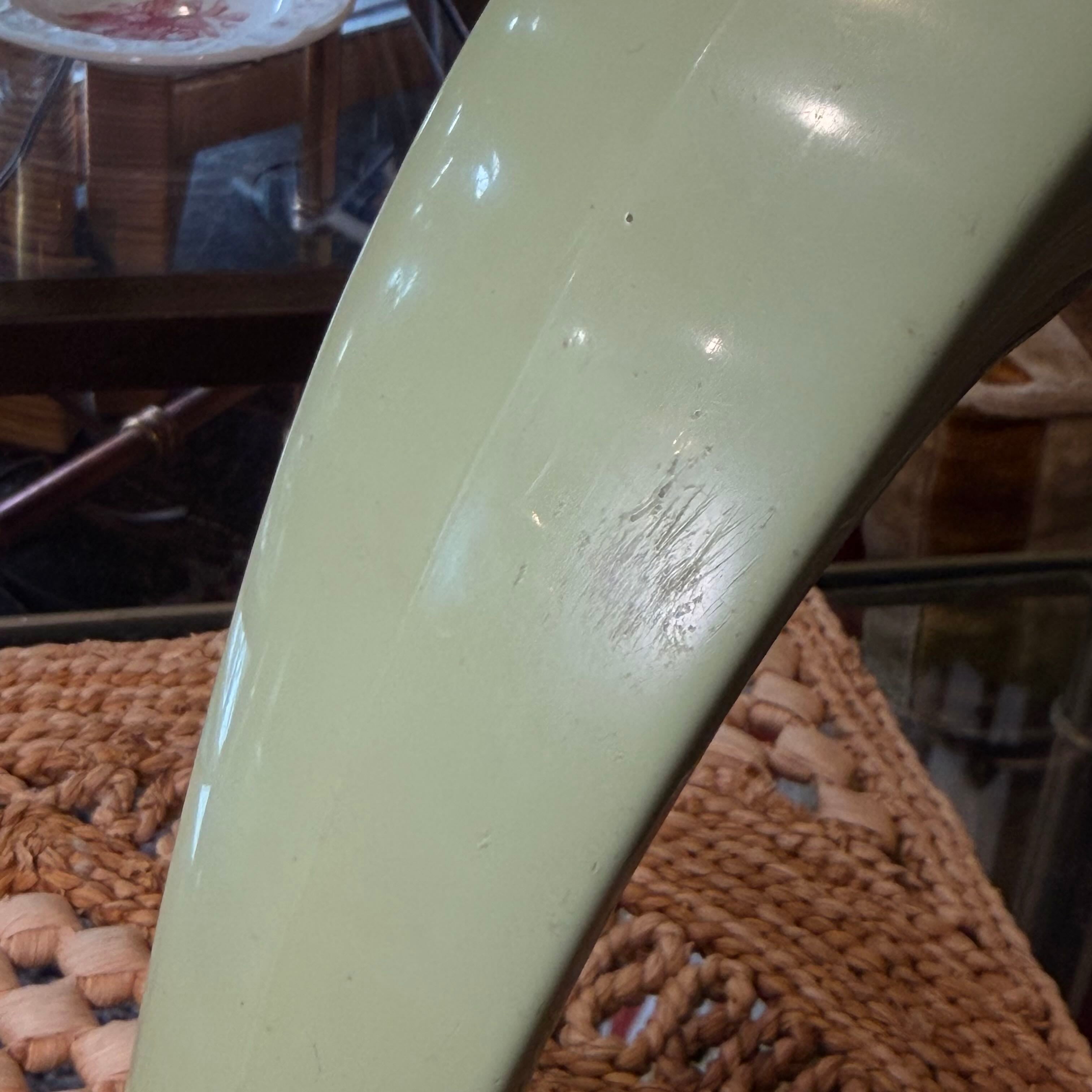 A close-up of a glossy pale sage green curved surface, likely part of a chair or furniture piece, resting on a woven brown textile with a blurred indoor background.