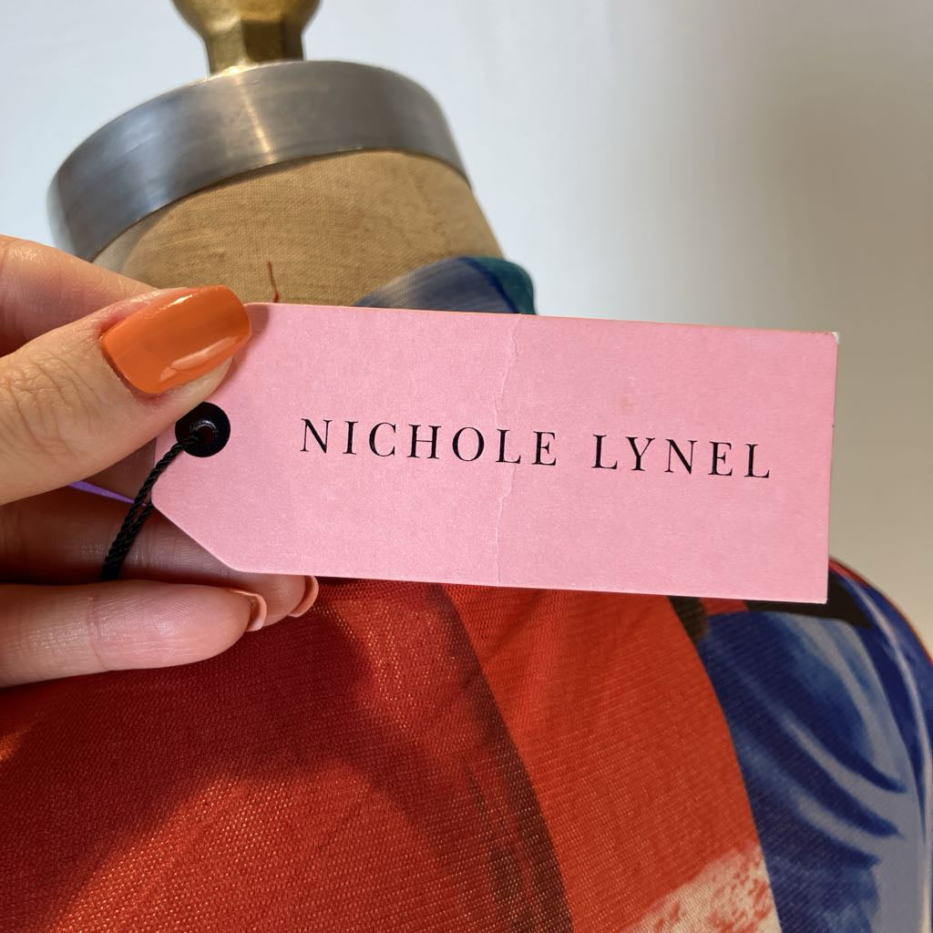 A hand with orange nail polish holds a pink paper brand tag reading 'NICHOLE LYNEL' in front of a mannequin wearing a colorful garment with orange, red, and blue tones.
