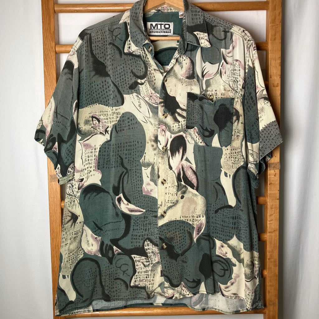 A short-sleeve button-up shirt with an abstract collage print featuring muted green, beige, and black tones, hanging on a wooden hanger against a white curtain backdrop.
