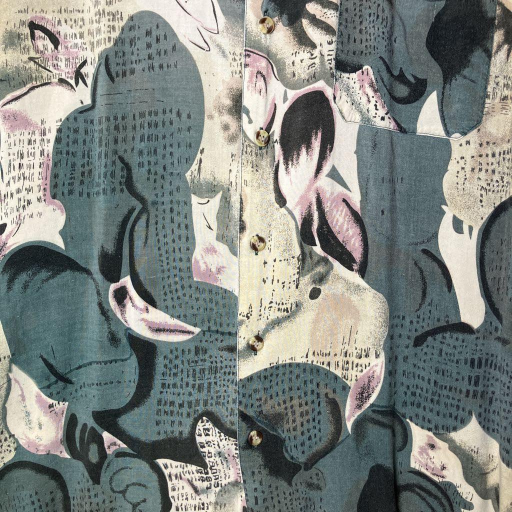 A close-up of a button-front garment showing an abstract collage with muted blue-gray, beige, black, and dusty pink tones.