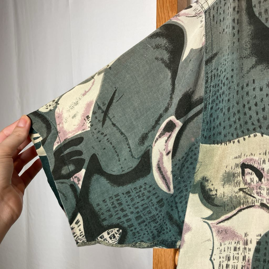 A close-up of a short-sleeve shirt being held by a hand, showing a pattern of muted teal, black, cream, and dusty pink tones.