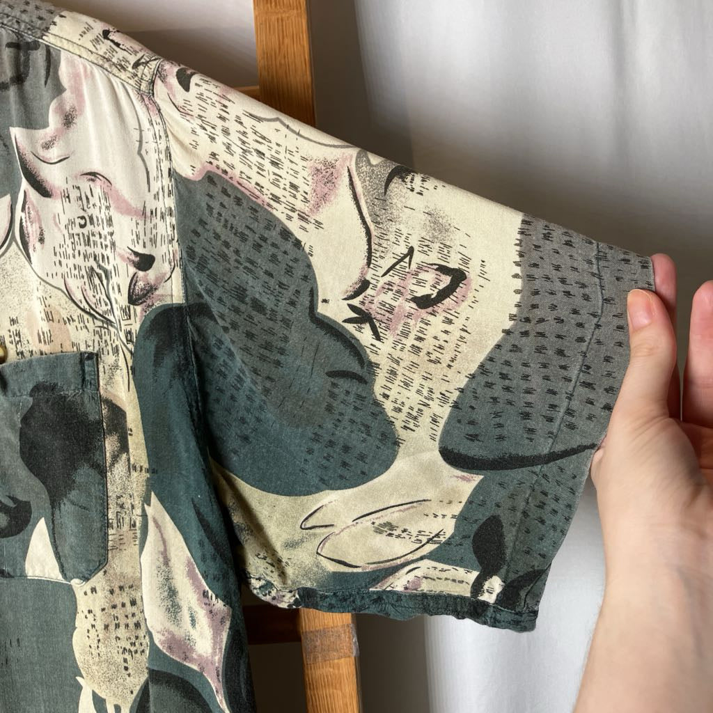 A close-up of a person's hand holding up a short-sleeve shirt or dress to show the fabric print. The garment features an abstract, painterly pattern in muted teal, beige, black, and soft pink, displayed on a wooden hanger against a white wall.