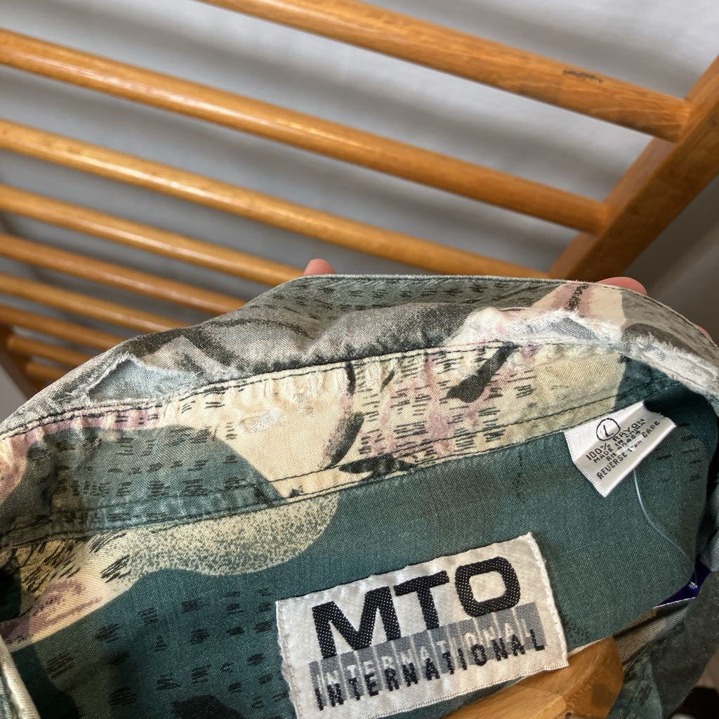 Close-up of the inside collar of a camouflage-patterned shirt or jacket, showing a sewn-on patch that reads 'MTO INTERNATIONAL' and a small white care label. A hand is holding the garment, with a wooden chair visible in the background.
