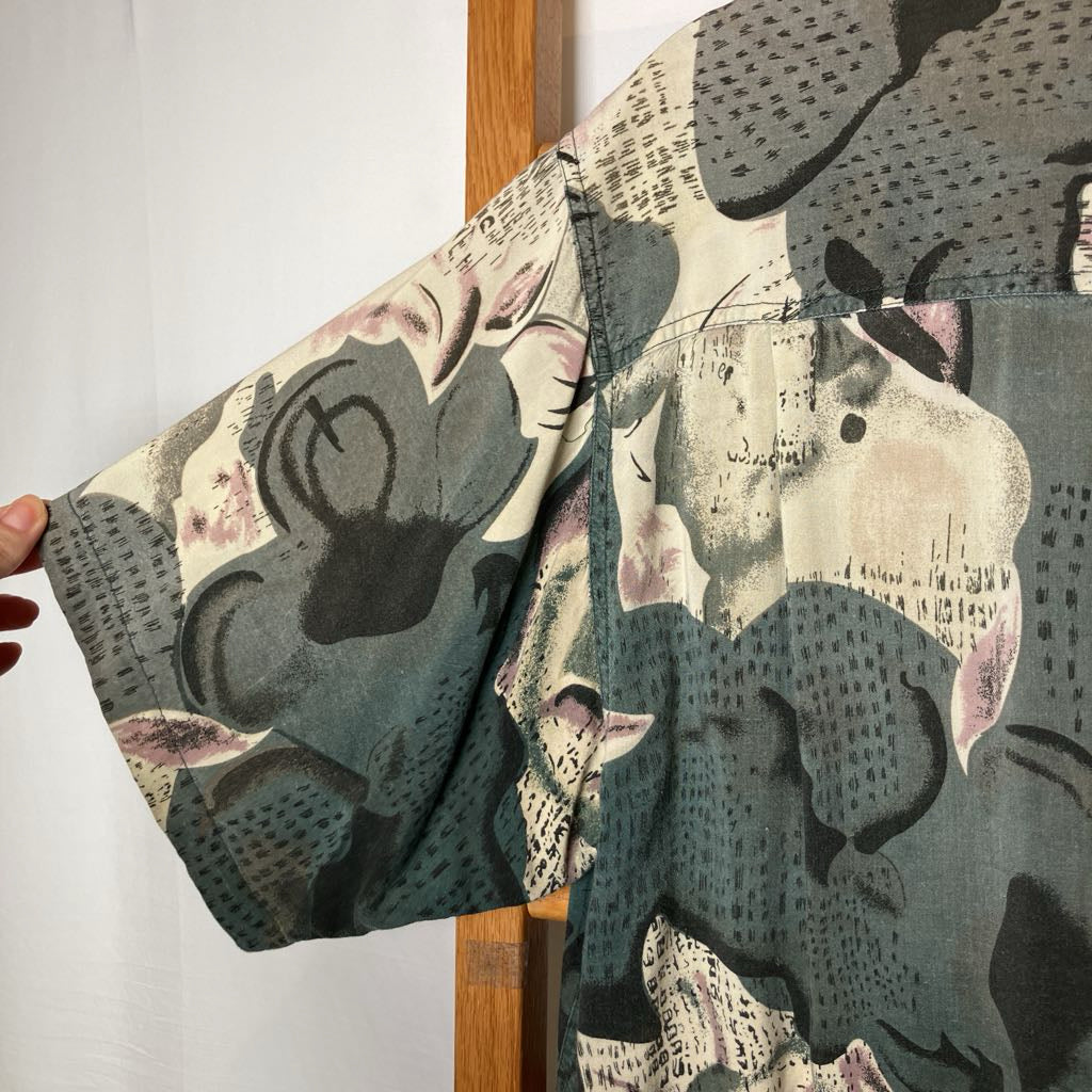 A close-up of a short-sleeve button-up shirt with a complex abstract collage print featuring muted gray, teal, beige, and pink tones. A hand is holding the sleeve to show its drape and width.