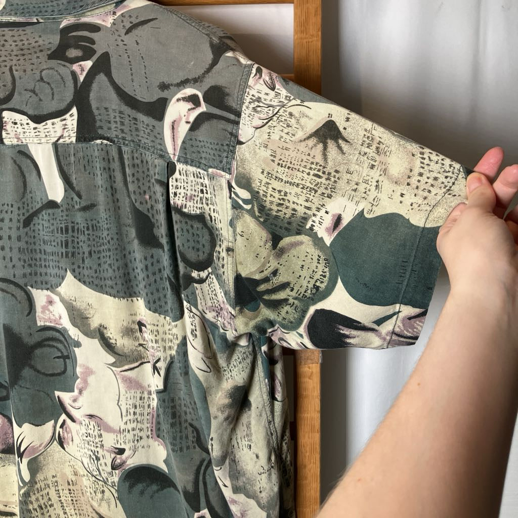 A close-up of a short-sleeve button-up shirt with a complex abstract collage print, held by a person's hand against a neutral background.
