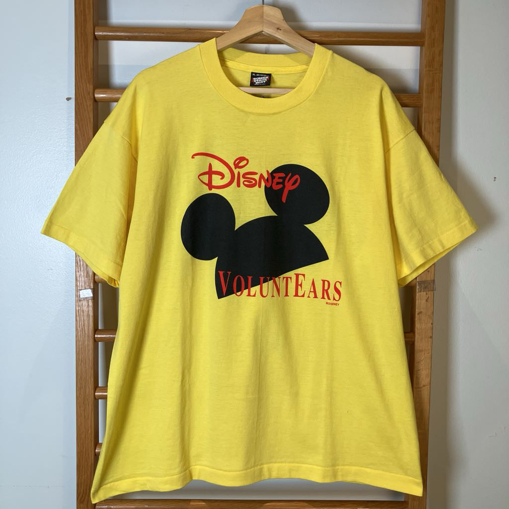 A bright yellow short-sleeve T-shirt hanging on a wooden hanger against a wooden ladder, featuring a large black Mickey Mouse silhouette with the red 'Disney' logo above and 'Volunteers' text below.