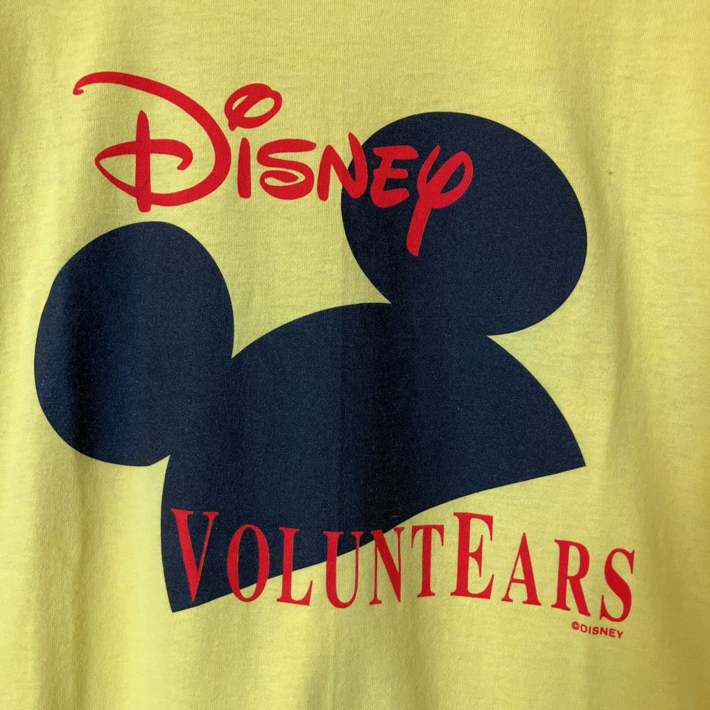 A close-up of a yellow T-shirt featuring a large navy blue Mickey Mouse silhouette graphic, with the red Disney logo above and the word 'VOLUNTEARS' printed below in red.