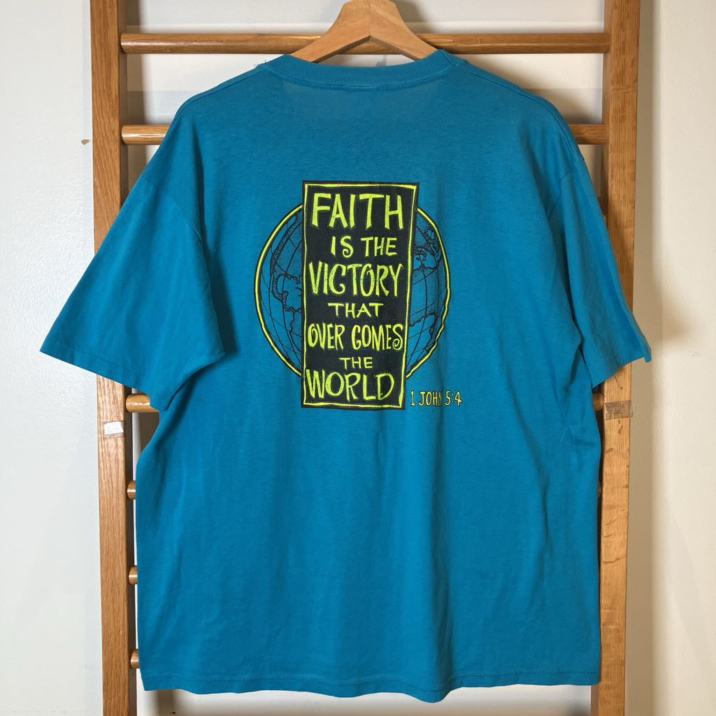 A teal blue t-shirt hanging on a wooden clothes hanger against a light-colored wall. The back of the shirt features a yellow graphic with the text 'FAITH IS THE VICTORY THAT OVER COMES THE WORLD' and a small '1 John 5:4' reference.
