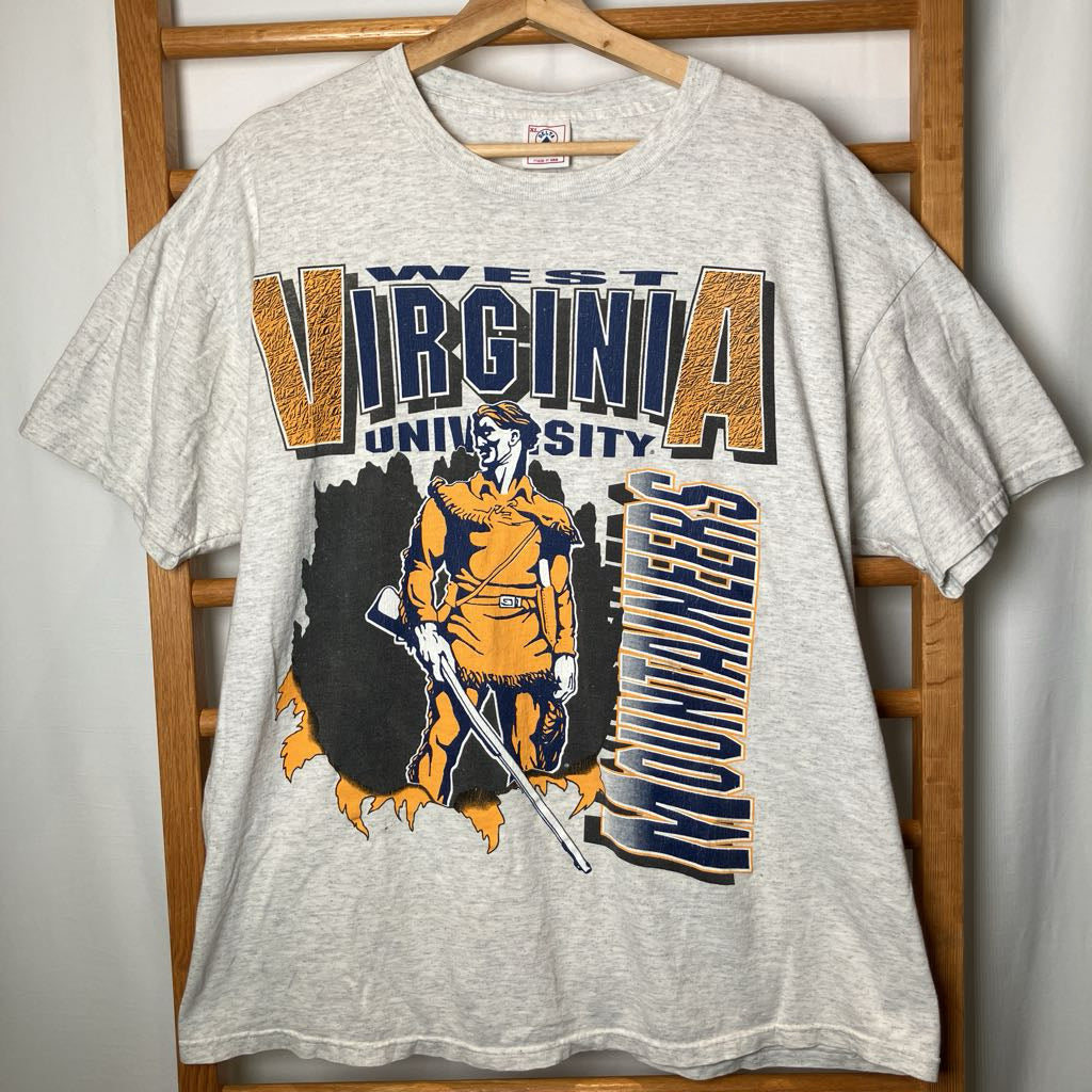 A heather gray vintage West Virginia University t-shirt hanging on a wooden hanger against a wooden chair. The shirt features a large graphic print with the team mascot, a sword, and the text 'WEST VIRGINIA UNIVERSITY' and 'MOUNTAINEERS' in orange, navy, and black.