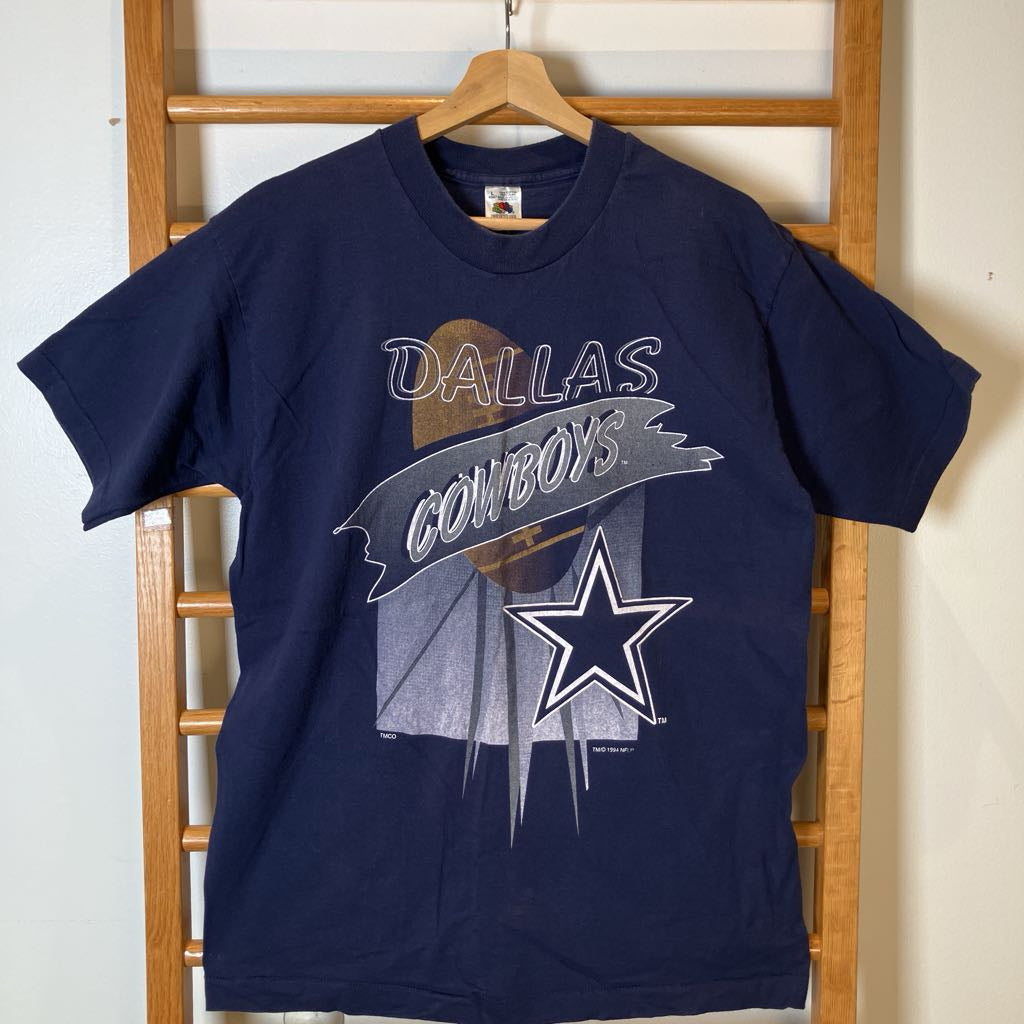 A navy blue Dallas Cowboys graphic t-shirt hanging on a wooden hanger from a wooden ladder rack against a light wall.