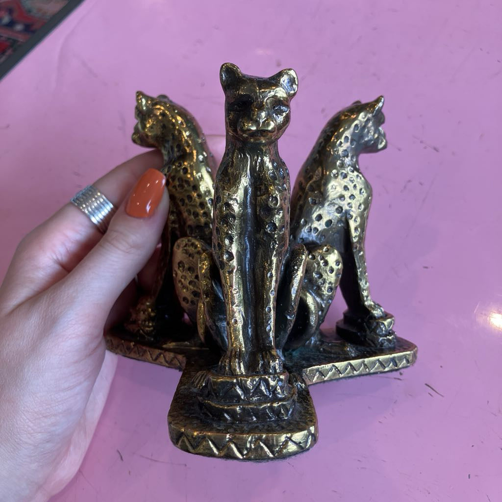 A hand with orange nail polish and a silver ring holds a small brass-finished decorative object featuring three spotted big cat figures on a pink background.