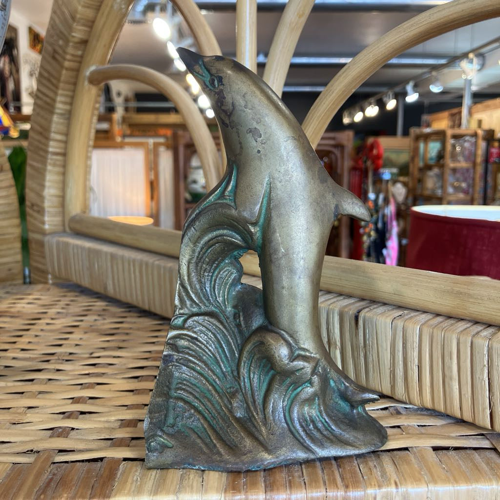 A vintage-style metal dolphin sculpture rising from stylized waves, displayed on a woven wicker surface in a shop setting.