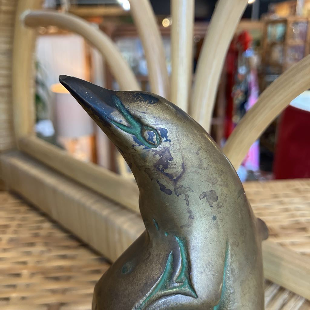 A close-up of a metallic bronze dolphin figurine with green patina accents, resting on a woven wicker surface with a blurred wooden chair in the background.