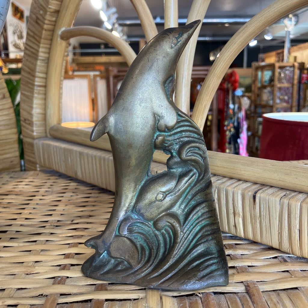 A bronze-finished metal dolphin sculpture standing on stylized wave bases, displayed on a woven wicker surface in a shop setting.