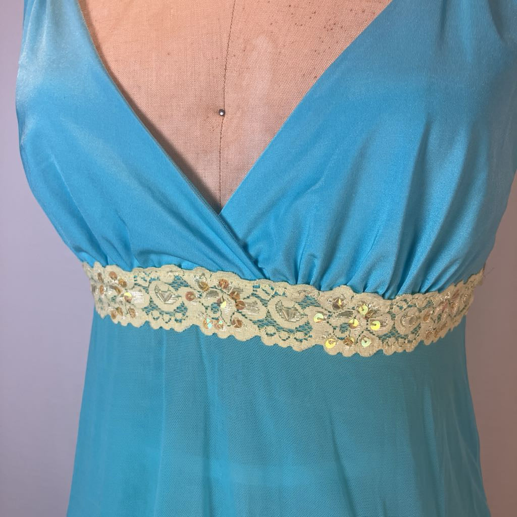 A close-up of a turquoise blue satin-like dress or nightgown displayed on a mannequin torso, featuring a deep V-neckline and a wide decorative waistband with beige floral lace and small gold sequins.