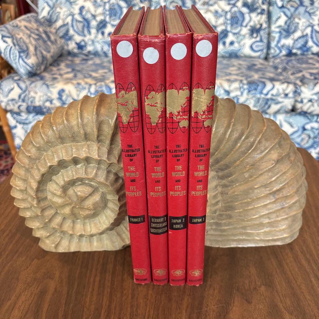 A set of four vintage hardcover books titled 'The Albeit Unwritten Library of the World and Its Peoples' by Kenneth Hahn, standing upright on a wooden table between two large beige shell-shaped bookends. The books have red covers with gold lettering and white circular stickers near the top.
